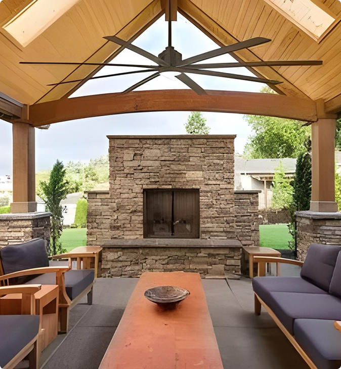 Outdoor patio with wooden furniture, a long table, and stone fireplace under a wooden pergola. A large ceiling fan is mounted above. Greenery visible in the background.