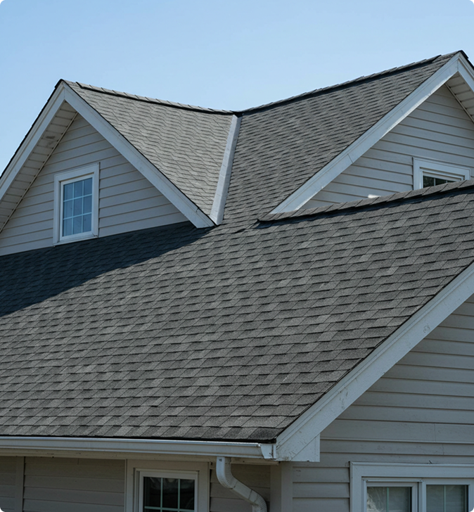 Gabled roof of a house with dark gray shingles, featuring two dormer windows and beige siding, under a clear blue sky.