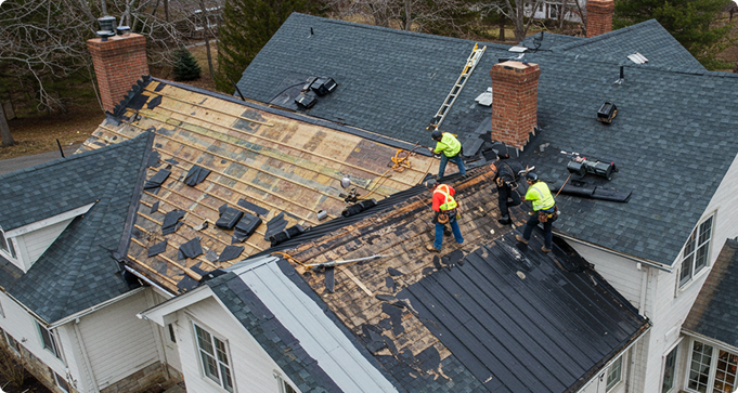 Roofers working on a large residential roof with missing shingles and visible wooden structures. Ladders and tools are present.